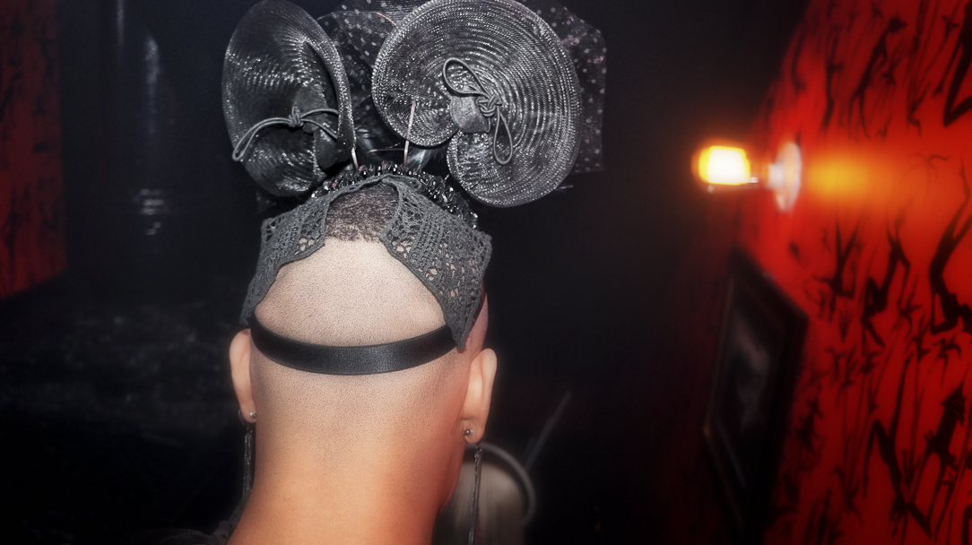 A sculptural headpiece photographed from behind in a red and black club interior