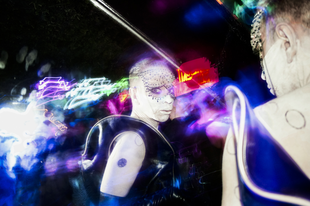 A clubgoer in black vinyl reflected in a mirror amid electric blue and magenta light trails