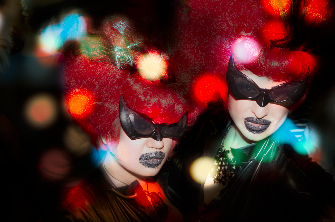 Two clubgoers in red sculptural wigs and dark glasses framed by colored light